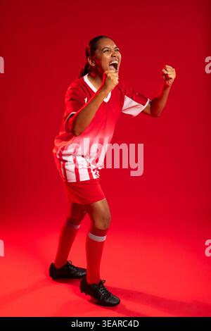 African American woman celebrating and clenching fists in red white jersey, shorts, socks at studio. Athlete, victory, energetic, dynamic, sportswear, Stock Photo