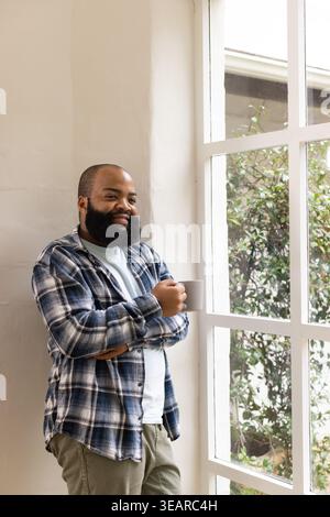 African American man standing at home by paned window holding ceramic mug, plaid shirt, copy space. Sunlit, serene, cozy, contemplative, bearded, mull Stock Photo