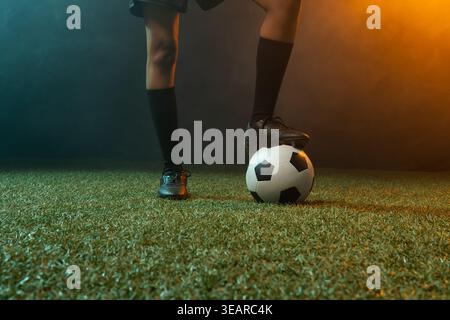 African American woman standing on turf balancing on soccer ball in cleats under colored lights. Athlete, balance, focus, intensity, lighting, haze, s Stock Photo