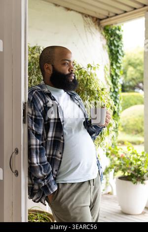 Adult African man wearing plaid shirt leaning in doorway, holding ceramic mug, gazing toward garden. Porch, veranda, outdoor, greenery, bearded, cup, Stock Photo