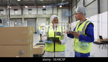Two workers checking inventory in depot, wearing vests and hairnets, holding tablet and clipboard. Warehouse, teamwork, pallets, crates, stock, logist Stock Photo