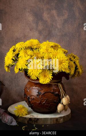 Vertical still life of yellow dandelions in a textured clay jug on a wooden slice. Rustic floral arrangement with a warm, dark brown background. Stock Photo