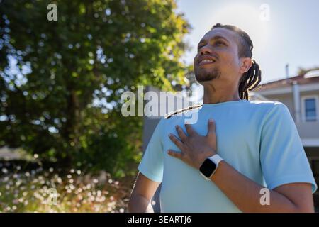 African American man standing hand on chest looking up yard wearing shirt smartwatch, copy space. Dreadlocks, adult, backlight, outdoor, garden, subur Stock Photo
