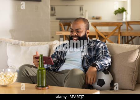 African American man sitting on light sofa at home holding smartphone and resting on soccer ball. Phone, livingroom, relaxed, cozy, lifestyle, home, o Stock Photo