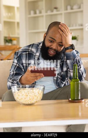 Adult African American man sitting on sofa holding red phone snacking on popcorn, bottle on table. Cozy, casual, warm, livingroom, decor, shelves, pla Stock Photo