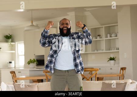 African American man wearing flannel shirt raising fists, cheering at home near sofa, green bottle. Celebration, exuberant, openplan, livingroom, natu Stock Photo