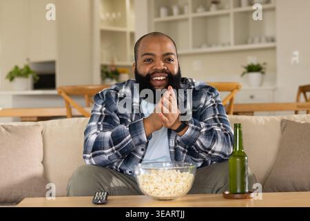 African American man in plaid flannel shirt clapping while watching TV in living room with popcorn. Bearded, cozy, home, couch, openplan, kitchen, amb Stock Photo