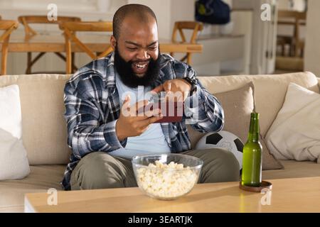 African American man sitting on sofa holding smartphone, popcorn bowl, beer bottle and soccer ball. Casual, cozy, home, livingroom, plaid, cheerful, r Stock Photo