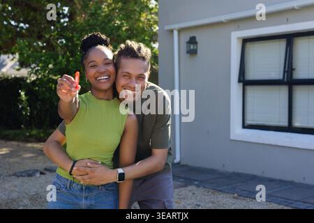African American couple smiling and holding single house key on red fob in front of house. Outdoor, frontyard, partners, homeownership, sunny, exterio Stock Photo