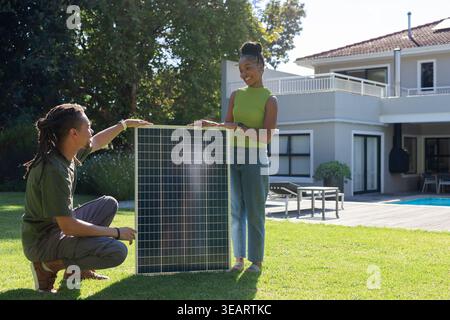 African American couple installing large photovoltaic solar panel on grassy lawn near pool. Backyard, deck, balcony, greenery, sunlit, energy, renewab Stock Photo