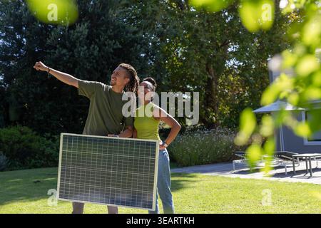 African American couple holding solar panel, pointing outward at home, woman in lime top, jeans. Photovoltaic, backyard, lawn, patio, sunlit, teamwork Stock Photo