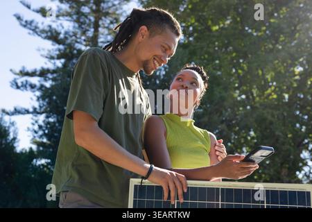 African American couple in summer tees leaning on PV panel checking meter showing wristband in yard. Solar, array, outdoor, sunlight, collaboration, s Stock Photo