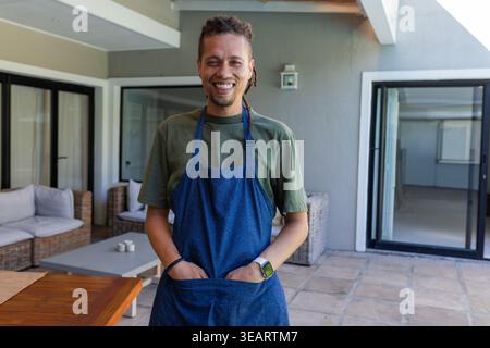 African American man standing and smiling on veranda wearing denim apron, hands tucked in pocket. Outdoor, patio, wicker, host, chef, smartwatch, casu Stock Photo