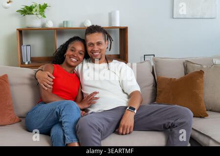 African American couple sitting close on light beige sofa, leaning in and smiling with smartwatch. Livingroom, partners, warmth, relaxed, cozy, neutra Stock Photo