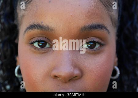 African woman looking directly at camera in close-up showing hoop earrings, mascara, skin. Portrait, headshot, gaze, makeup, jewelry, curls, hair Stock Photo