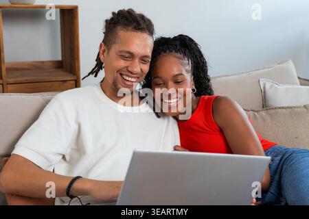 African American couple sitting on sofa at home, woman wearing red top, smiling at laptop. Duo, partners, device, domestic, living, room, cozy Stock Photo