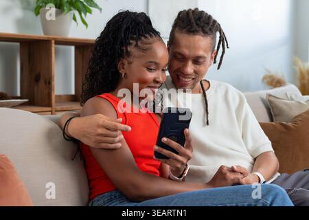 African American couple sitting on couch in living room, woman in red top looking at smartphone. Togetherness, cozy, naturallight, sofa, phone, plant, Stock Photo