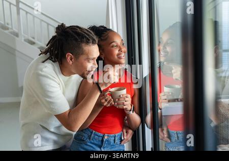 African American couple standing by glass door, man pointing outside, woman in red holding mug. Window, reflections, interior, airy, daylight, stairca Stock Photo