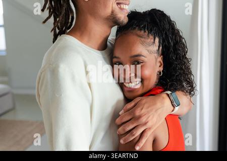 African American couple embracing in bright home, man with smartwatch, woman in red top and hoops. Affection, intimacy, partners, dreadlocks, curly, k Stock Photo