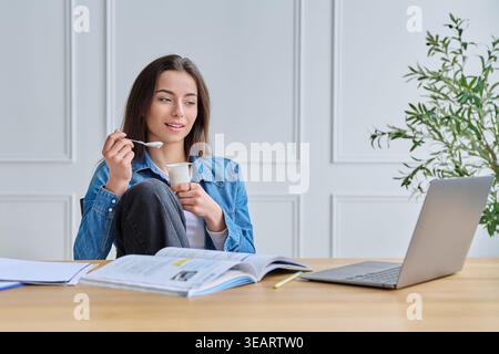 Young woman in denim shirt eating yogurt while studying with laptop and books Stock Photo