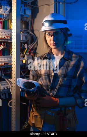Female electrical engineer standing near switchboard holding system blueprint. Professional portrait Stock Photo