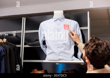 Male customer checking price on shirt while standing in clothing store, looking at red sale tag sticker. Man shopper counting discount while making purchases in clothes store on Black Friday Stock Photo