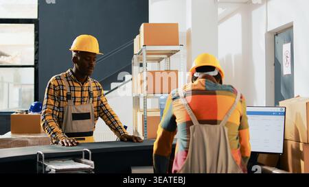 African american employees doing fist bump gesture in warehouse, greeting each other at work. Team of men working in storage room with containers and boxes of merchandise. Handheld shot. Stock Photo
