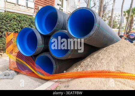 Large black corrugated plastic pipes with blue interior stacked on a construction site next to a pile of sand and orange safety net. Stock Photo