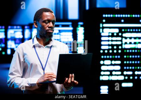 IT programmer in server hub using laptop to find network issues affecting supercomputer performance. Employee fixing issues leading to slow data transfer rates and poor client communication Stock Photo