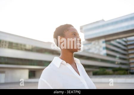 Smiling businesswoman with short hair looking away while standing near modern office buildings, sunlight on face with confident expression Stock Photo