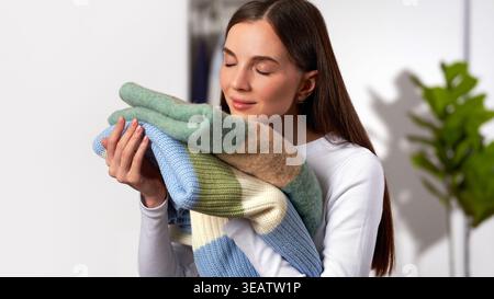 Young Caucasian white woman holding and smelling clean sweaters on light gray wardrobe in home background. Concept of fresh laundry, woolen clothing c Stock Photo