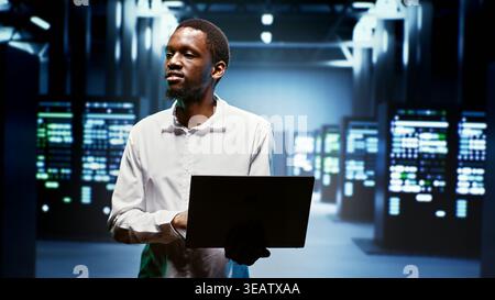 African american repairman using laptop to review energy consumption across server cabinets components. Manager making sure data center equipment temperature sensors are performing flawlessly Stock Photo