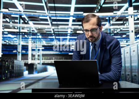 Overseeing supercomputers, laptop and technician in data center with large scale IT infrastructure. Doing network management virtualization, notebook and man working in server farm Stock Photo