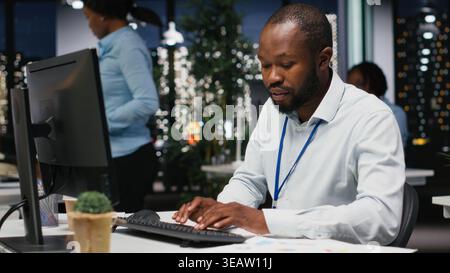 African american male worker proofreading financial numbers, reviewing archived files after hours in the strategy room. Man evaluating data on reports and doing research. Stock Photo
