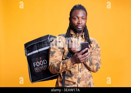 Pizzeria courier checking customers adreess on phone using takeaway food app before delivery food order. African american restaurant employee carrying thermal backpack standing in studio Stock Photo