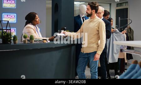 Shopper holds smartphone at pos terminal, making contactless payment with nfc technology while shopping for clothes. Man at cashier counter using mobile phone to pay for purchases. Stock Photo