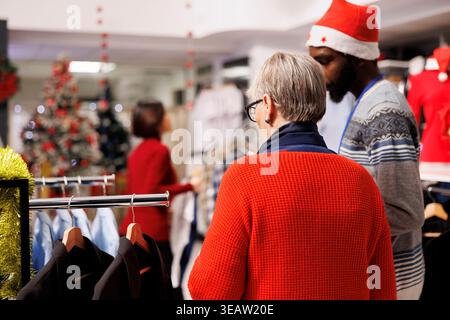 Retail worker helps senior woman with shopping during christmas season, presenting items on racks to choose as gifts. Employee recommending clothing for old person, seasonal sales. Stock Photo