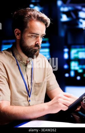 Repairman using tablet to examine server racks for power fluctuations, ensuring uninterrupted computing power. Worker monitoring data center, identifying potential vulnerabilities Stock Photo