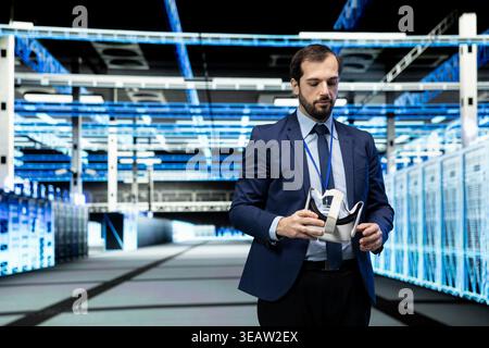 Data center developer putting VR goggles equipment on to configure server settings. Man in modern high tech server hub uses virtual reality simulation to do maintenance tasks on supercomputers Stock Photo