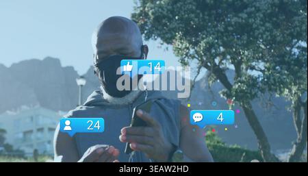 Scrolling man wearing sleeveless hoodie and black mask using phone at park, wired earphones, likes Stock Photo