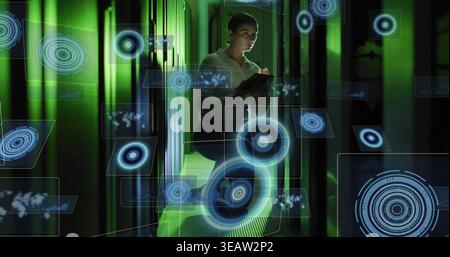 Inspecting female tech scanning server LEDs in datacenter holding tablet, in blouse viewing holo UI Stock Photo