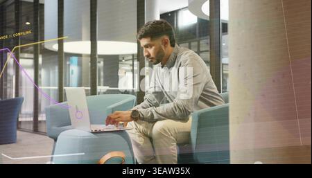 Typing Adult Indian man wearing gray shirt, khaki pants, using laptop on ottoman in office lounge Stock Photo