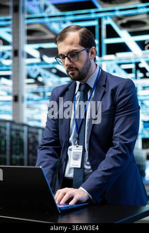 Data center programmer at desk configuring server settings with laptop software. Worker in modern high tech server farm using notebook to do maintenance tasks on IT infrastructure machines Stock Photo