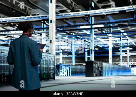Data center computer scientist coding on tablet, upgrading autonomous IT infrastructure rigs.IT expert walking in server farm, using programming script app on device to update systems Stock Photo