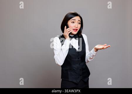 Catering service employee in uniform explaining to customer while answering call on smartphone. Young asian woman receptionist having conversation with hotel staff on mobile phone Stock Photo
