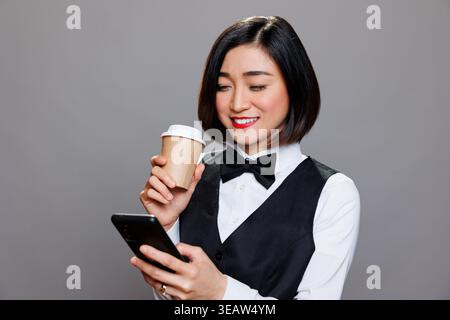 Smiling asian woman dressed in waitress uniform holding coffee paper mug and browsing internet on smartphone. Receptionist enjoying tea and engaging with social media networks on mobiel phone Stock Photo