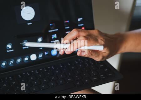 A close-up shot of a hand using a white digital stylus to navigate a tablet computer attached to a black keyboard case. Stock Photo