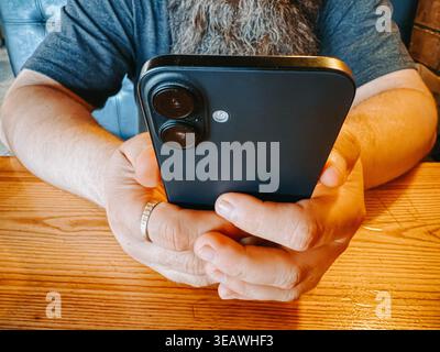 Valmiera, Latvia - April 22, 2026: Close-up of a bearded man holding a smartphone while sitting at a wooden table. Ideal for concepts like mobile comm Stock Photo