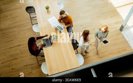 A diverse group brainstorms in an open office, sharing ideas and collaborating at a large wooden table under natural light. Stock Photo