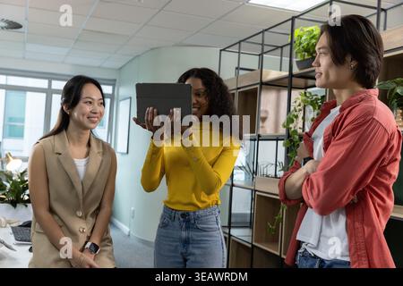Diverse coworkers standing together sharing black tablet, leaning on desk near laptop and plants Stock Photo